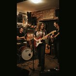 Fotografie zachicuje kapelu v zkušebně. V popředí stojí žena s červenou elektrickou kytarou, za ní muž za bicím sadou značky Gretsch a vpravo muž s č…