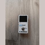 Bílý kytarový ladič s černým displejem. Na přední straně jsou nápisy „display“, „pitch“, „tuning“, „output“ a „input“. Uprostřed je modrý a černý náp…