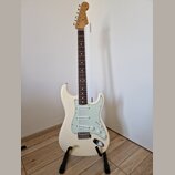 Bílá elektrická kytara s jasně viditelným logem Fender na hlavě. Nástroj má hnědý hmatník, světle zelený pickguard a tři snímače. Kytara je umístěna…