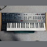 Široký analogový synthesizér s klaviaturaí a dřevěným bočním rámem. Horní panel je šedý s mnoha ovládacími knoflíky a přepínači, rozdělenými do sekcí…