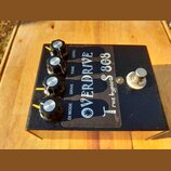 Černý kytarový efekt s nápisem OVERDRIVE TS 808 a doplňkem true bypass. Na horní straně jsou čtyři černé otočné knoflíky s kovovými vršky a popisky L…