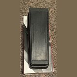 Černý sustain pedál s žebrovaným povrchem, umístěný na bílé krabici. Na pedálu jsou vyrazeny nápisy INSTRUMENT a AMPLIFIER. Povrch plastu vykazuje mí…