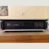 Černý výkonový zesilovač v hliníkovém racku. Na předním panelu je bílý nápis QSC AUDIO a model RMX 1850HD PROFESSIONAL POWER AMPLIFIER. Vpravo jsou o…