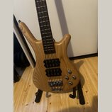 Světle hnědá elektrická basová kytara s přírodným dřevem a viditelnou masivní strukturou zrna. Nástroj má dva černé humbuckery, čtyři ovládací knoflí…