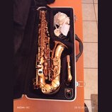 V otevřeném černém kufru leží saxofon s lesklým, růžozlatým povrchem. Vedle nástroje je umístěn mouthpiece s troškou rýžového rýdu a bílá látka. Na n…
