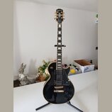 Černá elektrická kytara typu Les Paul s bílými hrankami a zlatým hardwarem. Na hlavě je nápis Epiphone a logo v podobě křídla. Kytara má dva humbucke…
