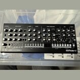 Černý syntezátor s nápisem SE-02 STUDIO ELECTRONICS a logem Roland. Přední panel obsahuje množství otočných potenciometrů, tlačítek a přepínačů rozdě…