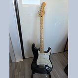 Černá elektrická kytara s bílým pickguardem a hnedým javorovým hridlem. Na hlavě je viditelné logo Fender a nápis Stratocaster. Nástroj vykazuje znám…