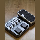 Šedý kytarový procesor s ovládacími knoflíky, displejem a dvěma černými spínacími pedály. Na přístroji jsou viditelné nápisy MG-100, Modeling Guitar…