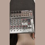 Stříbrný mixážní pult s nápisem BEHRINGER XENYX 1202FX. Zařízení obsahuje několik vstupních kanálů s potenciometry, přepínači a konektory typu XLR a…
