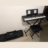 Černý digitální klavír umístěný na černém skládacím stojanu s přívodním kabelem. Na nástroji je vidět nápis YAMAHA a doprovodná hudební notová opěrka…