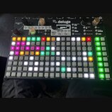 Černý sekvenční sampler s nápisem deluge. Zařízení má mřížku podsvícených tlačítek v různých barvách, několik otočných knoflíků a displej s nápisem S…