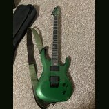 Zelená elektrická kytara s kovovými strunami a dvěma černými humbuckery. Na hlavě je viditelné logo Ibanez. Kytara má zelený popruh a vedle ní leží č…