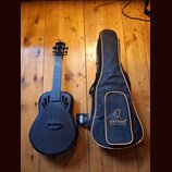 Černá kytara s povrchem imitujícím karbonové vlákno a doprovodný černý pouzdro s oranžovým lemováním. Na pouzdře je bílé logo a nápis ORTEGA GUITARS…