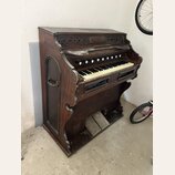 Hnědý dřevěný pomníkový orgán s klaviaturaí. V horní části je vyřezávaný ornament a nápis "SAYER & SON". Povrch dřeva vykazuje známky opotřebení a od…