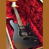 Černá elektrická kytara s matným povrchem v červeném plišovém kufru. Nástroj má dva humbuckery, černý pickguard a světlou hmatnici. Na hlavě kytary j…