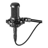 Černý kondenzátorový mikrofon umístěný v odpruženém držáku na černém stativu. Na těle mikrofonu je bílý nápis audio-technica. Na spojovací části stat…
