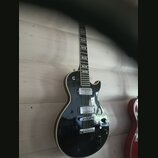 Černá elektrická kytara s tvarem single-cutaway a bílým lemováním kolem korpusu. Nástroj má dva humbuckery, Tune-o-matic můstek a čtyři ovládací knof…