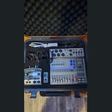V černém přepravním kufru s pěnovou výplní se nachází digitální mixáž ZOOM LiveTrak L-8, modul Roland TR-505 a bílý napájecí adaptér s kabelem. Na mi…