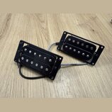 Dvě černé humbuckery s montážními rámečky, položené na dřevěném povrchu. Snímače mají viditelné kovové šroubky a pólové piny. Na fotce nejsou žádné č…
