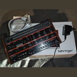 Černý syntezátor s červenými ovládacími prvky a nápisy. Na přístroji je viditelné logo behringer a název modelu GRIND. Vedle nástroje leží bílá krabi…