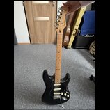 Černá elektrická kytara s bílým pickguardem a javorovým hnedým hmatníkem. Na hlavě je viditelné logo Fender a nápis STRATOCASTER. Kytara má tři sníma…