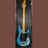 Modrá elektrická basová kytara v černém tvrdém kufru s nápisem MUSIC MAN. Nástroj má světle hnědý hриф z dřeva a černý pickguard. Na hlavě je viditel…