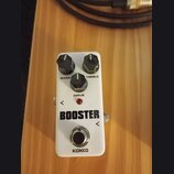 Bílý kytarový efekt s nápisem BOOSTER a značkou KOKKO. Na horní straně jsou tři černé otočné knoflíky s popisky BASS, DRIVE a TREBLE. Uprostřed se na…