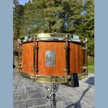 Dřevěný buben s přirozeným povrchem a zlatými kovovými obručemi. Uprostřed korpusu je kovový štítek s logem a nápisem CUSTOM DRUMS. Nástroj vykazuje…