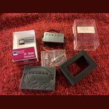 Na červeném povrchu je položen kytarový snímač typu humbucker v kovovém krytu s černými vodiči. Vedle něj je bílá krabička a červený papírový obal s…