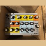Stříbrný analogový syntetizátor Moog Sirin umístěný v kartónovém boxu. Přední panel obsahuje množství ovládacích knoflíků rozdělených do barevných se…