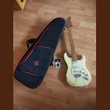 Bílá elektrická kytara s jasným hnedým hřidnicem a bílým pickguardem. Na hlavě je viditelné logo Fender. Vedle kytary leží černý obal s růžovým lemov…