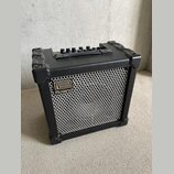 Černý kompaktní kombo zesilovač s kovovou mřížkou reproduktoru. Na přední straně je umístěno logo a nápis Roland. Horní panel obsahuje ovládací knofl…