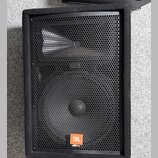 Černý reproduktor v bedně potažené látkou s kovovou mřížkou. V dolní části je oranžový čtverečkový štítek s bílým nápisem JBL a pod ním menší text JR…