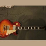 Elektrická kytara s tvarem singlecut a povrchovou úpravou v červeně-žlutém gradientu (sunburst). Má dva humbuckery, bílý pickguard a čtyři ovládací k…