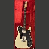 Kytara v krémové barvě s černým plechtrádem, položená v červeném pouzdře. Na hlavě hnedku je vidět nápis Squier by Fender a Telecaster. Nástroj má dv…