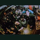 Fotografie zachycuje koncertní scénu s davem lidí v uzavřeném prostoru. V popředí je skupina osob v tmavém oblečení, z nichž někteří gestikulují. V p…