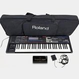 Černý syntetizátor s klaviaturou, na kterém je vpravo nápis Roland JUNO-60. Součástí sady je černý transportní vak s bílým logem Roland, napájecí ada…