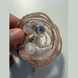 Sluchátka in-ear s průhledným plastovým tělem a měděně zbarveným pleteným kabelem. Na vnitřní straně jednoho sluchátka je viditelné černé kulaté logo…