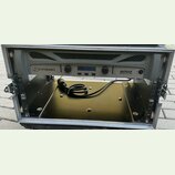 Šedý rackový zesilovač s nápisy crown a 4002 XTi SERIES. Zařízení má dva otočné ovladače, displej a několik tlačítek. Je umístěno v transportním kufr…