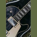 Detailní záběr na černou elektrickou kytaru v otevřeném kufru. Nástroj má hnědý hmatník s bílými blokovými vložkami a krémový pickguard. Na těle jsou…
