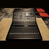 Černý digitální mixážní pult s displejem a několika řadami posuvníků. Na horním panelu jsou umístěny různé ovládací knofíky, tlačítka a digitální dis…