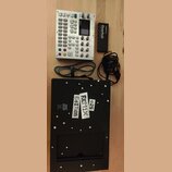 Na fotografii je stříbrný sekvencer Digitone s displejem a řadou tlačítek a otočných ovladačů. Vedle něj je černý adaptér s nápisem Overhub a dva nap…