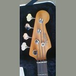Hlava čtyřstrunné basové kytary z přírodního dřeva v černém kufru. Na hlavě je vidět logo Fender a nápis PRECISION BASS, pod kterým je drobný nečitel…