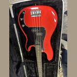 Červená elektrická basová kytara v černém pouzdře. Nástroj má černý pickguard a dvě kontrolní knoflíky. Na kytaru nejsou viditelné žádné značky ani n…