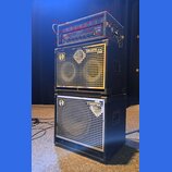 Svrhní část tvoří zesilovač s nápisem Mesa Boogie a červeným štítkem Max Carbine, který je přeškrtnutý červenými čarami. Pod ním jsou dvě basové repr…