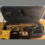 Zlatý saxofon v černém kufru. Vedle nástroje jsou položeny žlutý látkový sáček s dvěma rýdy a krabička rýd s logem „G“ a nápisem „GENUINE“. Na krabič…