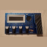 Modrý kytarový syntetizátor s nápisem Roland GR-55 GUITAR SYNTHESIZER. Zařízení má displej, ovládací knoflík se šipkami a čtyřistupňové pedály označe…