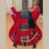Červená polodupatá elektrická kytara s černým plechtrátem. Nástroj má dva humbuckery, dva f-otvory a vibrační systém s páčkou. Na těle nejsou viditel…