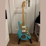 Modrá elektrická kytara s jasným dřevěným hřidlem a zlatým pickguardem. Na hlavě je viditelné logo "Supro". Kytara má dva humbuckery, dvě ovládací kn…